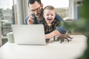 Vater mit Kind auf dem Schoß am Notebook und Handy als Metapher für "Mit Stress im Homeoffice entspannt umgehen"