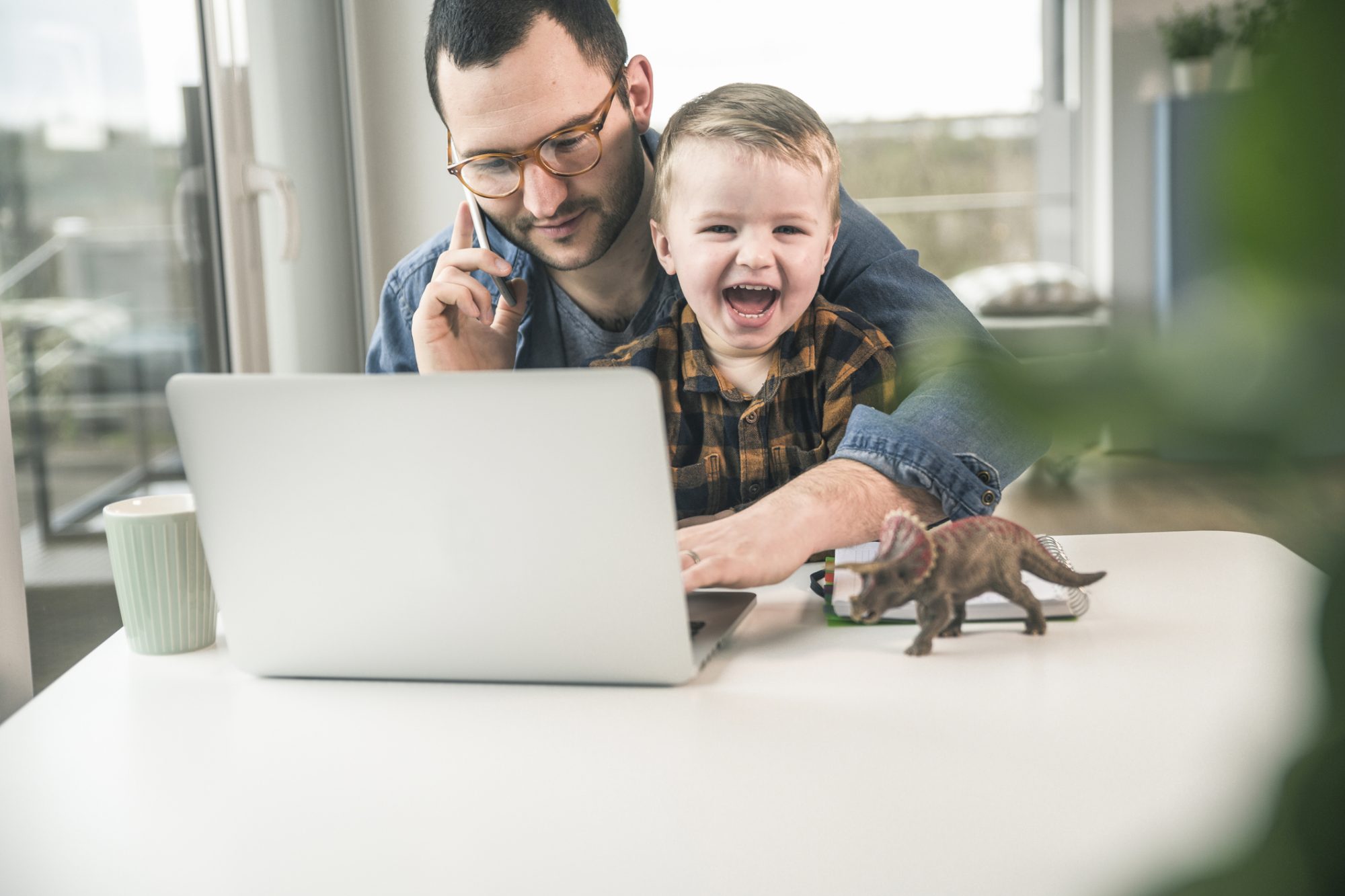 Vater mit Kind auf dem Schoß am Notebook und Handy als Metapher für "Mit Stress im Homeoffice entspannt umgehen"